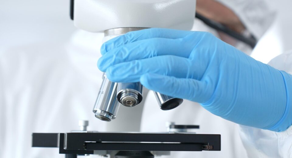 Close up of virologist's hands in blue gloves adjusting microscope in laboratory, conducting scientific research and analysis. Health care, medicine and science concept.